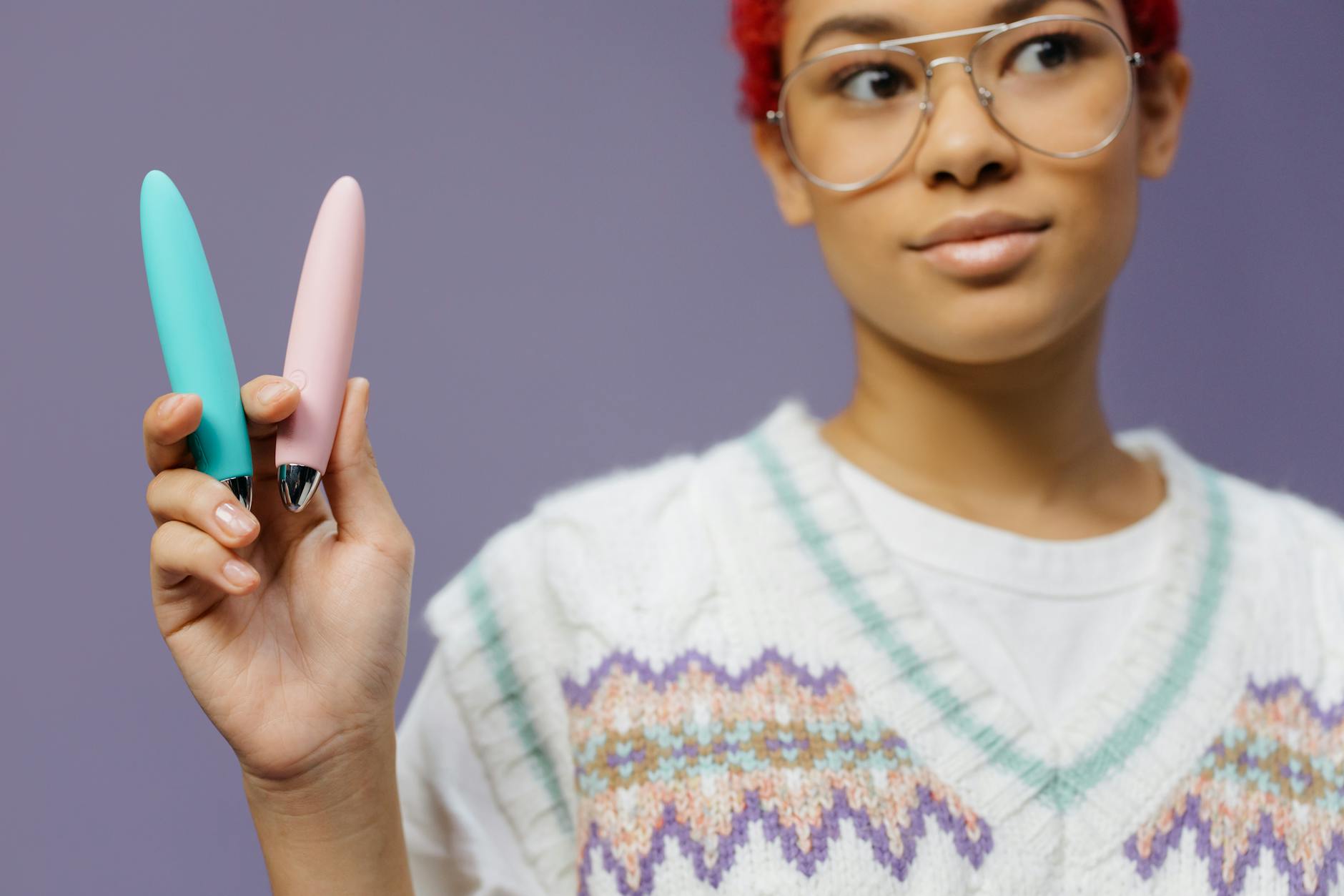 A woman with eyeglasses holds two colorful vibrators against a purple background.